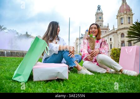 Zwei junge Frauen mit Einkaufstaschen und Blumen sitzen auf dem Gras vor dem Rathaus von valencia Stockfoto