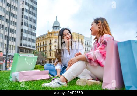 Zwei asiatische Frauen, die sich mit Einkaufstaschen auf dem Gras entspannen und sich nach einem erfolgreichen Shopping-Trip in der Stadt unterhalten Stockfoto
