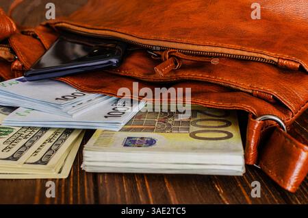 Nahaufnahme einer offenen braunen Ledertasche, die auf einer dunklen Holzoberfläche liegt und deren Inhalt enthüllt: Bargeldstapel einschließlich Euro (EUR) und US-Dollar Stockfoto