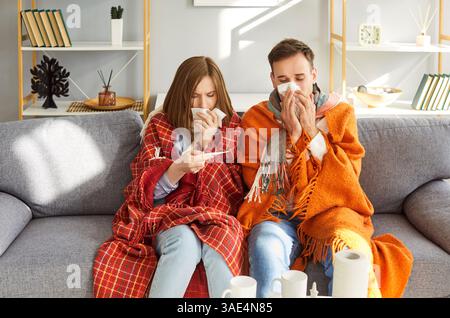 Junge ungesunde Paare sitzen traurig zusammen mit Decke auf Sofa und messen Temperatur Stockfoto