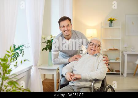 Junger glücklicher Sozialarbeiter, der mit älteren behinderten Menschen im Rollstuhl steht. Stockfoto