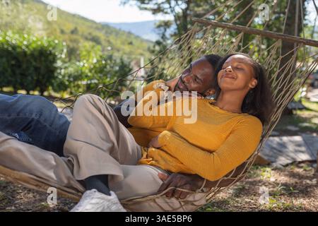 Sorglos lächelndes afroamerikanisches Paar, das seinen Urlaubstag in einer Hängematte verbringt und eine wunderschöne Glamping-Umgebung genießt. Stockfoto
