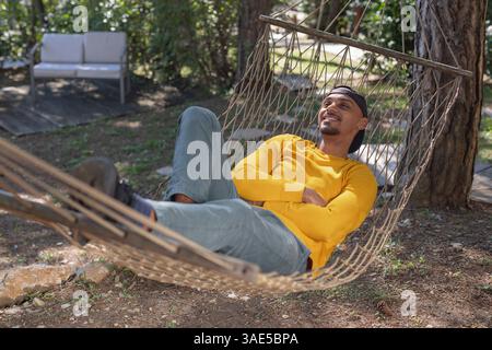 Junger afroamerikanischer Mann, der die Ruhe der Natur genießt, sich in einer Hängematte bewegt und entspannt. Wochenendausflug und Glamping-Konzepte. Stockfoto