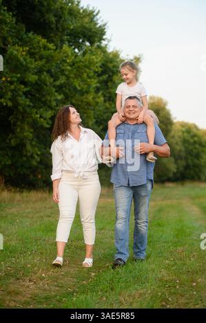 Familienspaziergang im Park. Dad trägt das Mädchen am Hals, Mom geht neben ihm und lächelt glücklich. Die Familie hat Spaß zusammen und läuft entlang p Stockfoto