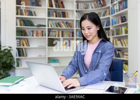 Eine junge Frau mit langen schwarzen Haaren arbeitet an ihrem Laptop in einer Bibliothek, Bücher im Hintergrund. Sie lächelt, während sie am Computer arbeitet. Stockfoto