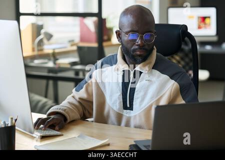 Junger afroamerikanischer Mann, der auf den Laptop-Bildschirm und die Tastatur des Desktop-Computers blickt, während er im Büro Netzwerkarbeit macht Stockfoto