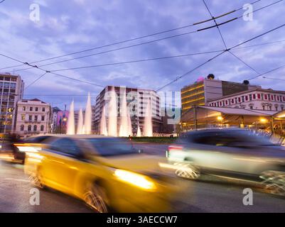 Athen, Griechenland - 5. April 2025: Omonoia-Platz in der Abenddämmerung mit verschwommenen Autos, beleuchteten Gebäuden und einem Brunnen im Hintergrund Stockfoto