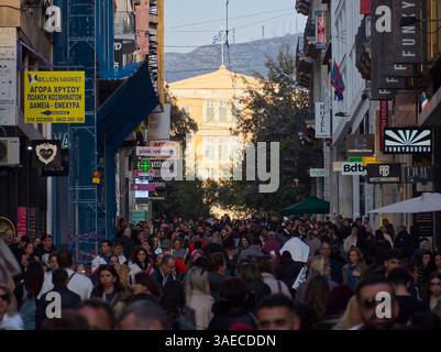Athen, Griechenland - 5. April 2025: Überfüllte Ermou-Straße mit Geschäften und Schildern auf Griechisch, Syntagma und Bergen im Hintergrund sichtbar Stockfoto