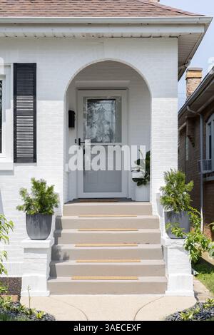 Eine Eingangstür mit Betontreppen führt zu einer bogenüberdachten Veranda, weißen Backsteinverkleidungen und schwarzen Fensterläden. Stockfoto