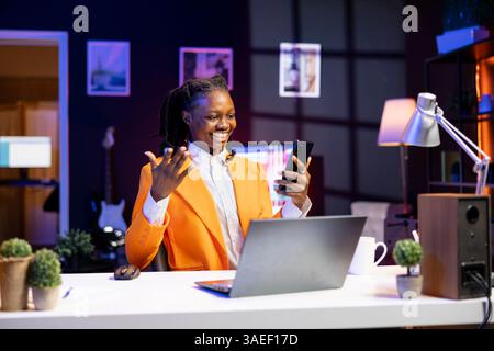 afroamerikanerin, die sich in der Wohnung entspannt, Freunde auf dem Handy schickt, Freizeit genießt. Lächelnder Remote-Mitarbeiter, der eine Pause einlegt, um mit Freunden im Internet über das Telefon zu sprechen Stockfoto