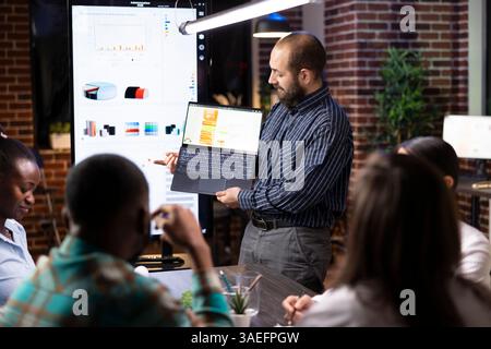 Kaukasischer männlicher Unternehmensberater präsentiert Geschäftsanalysen auf Laptop während des späten Nachtsitzens. Verschiedene Kollegen analysieren Finanzberichte und Brainstorming-Strategien im Startup Office. Stockfoto