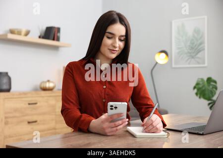 Schöne Frau mit Planer, die Notizen macht und Smartphone am Holztisch im Haus benutzt Stockfoto