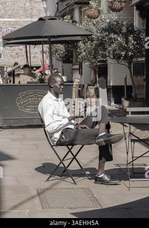 Windsor, Großbritannien - 03. April 2025 - Ein junger afrikanischer Mann trägt ein weißes Hemd, eine schwarze Hose und Lederschuhe, sitzt auf dem Stuhl und sieht sich Nachrichten auf dem Smartphone an Stockfoto