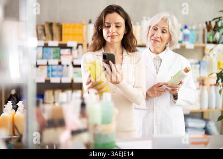 Frau in der Apotheke scannt QR-Code auf Shampoo, ältere Frau Apothekerin im Hintergrund Stockfoto
