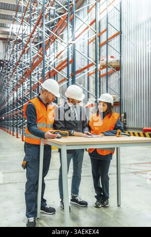 Lagerarbeiter und Manager, die Sicherheitshelme tragen, besprechen Logistikoperationen mit einem Barcode-Scanner und Dokumenten auf einem Tisch Stockfoto