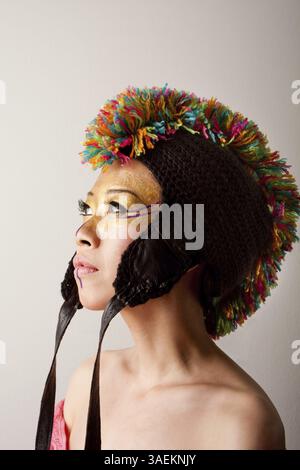 Schönes asiatisches Frauengesicht mit bunter mohawk-Hut mit Bändern, goldenem und lila Make-up, isoliert, New York, USA, Nordamerika Stockfoto