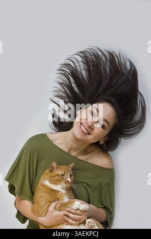 Eine Frau mit großem Lächeln in grünem Hemd, die ihr dunkelbraunes Haar schwingt, während sie eine orangene Katze hält, isoliert auf weiß, New York, USA, Nordamerika Stockfoto
