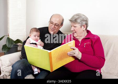 Stolze und glückliche Großeltern lesen Buch, um ihr Enkelkind zu erziehen. Baby-Mädchen, das Buch genießt, während es auf der Couch im Wohnzimmer sitzt, New York, USA, Nein Stockfoto