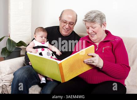 Stolze und glückliche Großeltern lesen Buch, um ihr Enkelkind zu erziehen. Baby-Mädchen, das Buch genießt, während es auf der Couch im Wohnzimmer sitzt, New York, USA, Nein Stockfoto