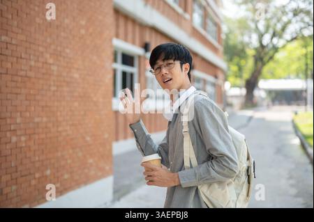 Ein asiatischer Mann mit Gläsern hält eine Kaffeetasse und winkt mit der Hand, während er in der Nähe einer Mauer steht. Gehen Sie im Freien oder gehen Sie irgendwo hin. Stockfoto