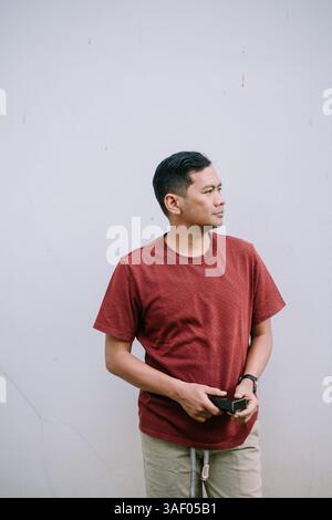 Ein stilvoller Mann in einem ziegelroten T-Shirt steht nachdenklich vor einem strukturierten Hintergrund. Mit echten Emotionen und natürlichem Licht aufgenommen Stockfoto