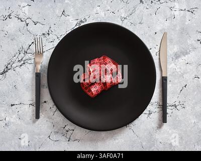 Rotes Maßband, das auf leerem schwarzen Teller mit Messer und Gabel auf Marmortisch liegt, repräsentiert Ernährung, Gewichtsverlust und gesunde Ernährung Stockfoto