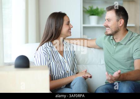 Glückliches Paar, das neben einem intelligenten Lautsprecher auf einer Couch zu Hause spricht Stockfoto
