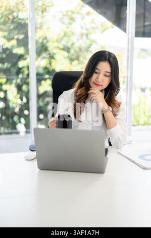 Eine asiatische Geschäftsfrau oder Büroangestellte, die eine Hand unter ihr Kinn hält und eine Kaffeetasse hält, während sie auf dem Schreibtisch glücklich auf den Laptop blickt. Stockfoto