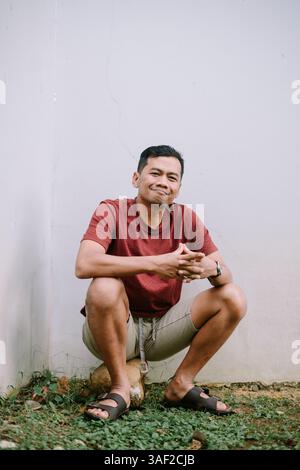 Porträt eines lächelnden und entspannten asiatischen Mannes, der vor der Wand sitzt und Frieden im Hinterhof seines Hauses ausdrückt Stockfoto