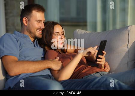Glückliches Paar, das das Handy benutzt und auf einem Sofa auf einer Terrasse sitzt, zu Hause oder im Hotel bei Sonnenuntergang Stockfoto