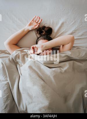 Draufsicht auf müde Frau im Bett mit Arm über Kopf und bedeckenden Augen. Stockfoto
