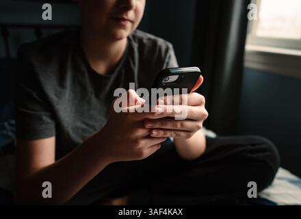 Nahaufnahme der Hände eines Teenager-Jungen mit Smartphone, der auf dem Bett sitzt. Stockfoto