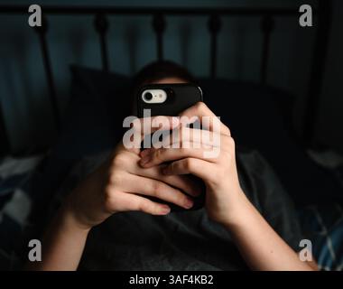 Nahaufnahme der Hände eines Teenager-Jungen, der ein Smartphone hält, das auf dem Bett liegt. Stockfoto