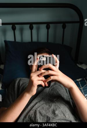 Nahaufnahme der Hände eines Teenager-Jungen, der ein Smartphone hält, das auf dem Bett liegt. Stockfoto