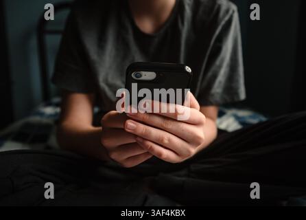 Nahaufnahme der Hände eines Teenager-Jungen mit Smartphone, der auf dem Bett sitzt. Stockfoto