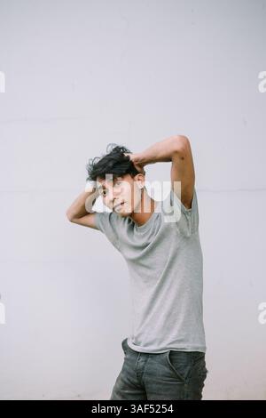 Ein junger Mann posiert vor einem sauberen, neutralen Hintergrund. Seine lässige Kleidung und sein selbstbewusster Ausdruck fesseln eine moderne, Jugendliche Energie. Stockfoto