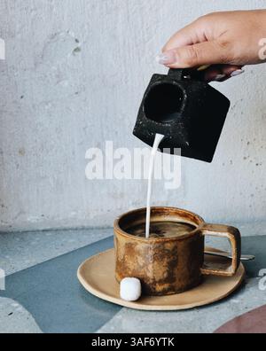 Tasse Kaffee, Becher und Teller auf brauner, moderner Keramik, Zuckerwürfel und eine Tasse Milch darüber. Blauer und grauer Hintergrund, saubere Ästhetik Stockfoto