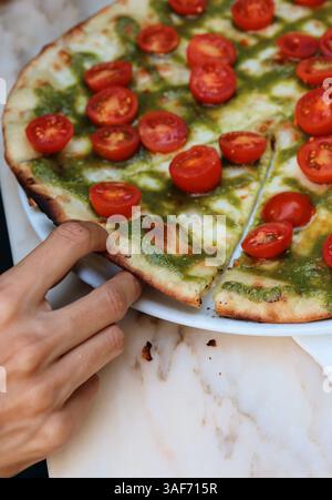 Dünne Hand, die ein Stück Pizza mit Hälften Kirschtomaten, Pesto und Käse auf eine Marmorplatte zieht Stockfoto