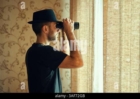 Mann mit Fedora-Hut, der durch ein Fernglas schaut und vom Fenster aus ausspioniert. Spion- und Suchkonzept Stockfoto