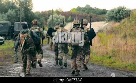 Soldaten marschieren mit militärischer Ausrüstung in Formation. Soldaten in Militäruniform mit Maschinengewehren laufen auf einer steinigen Straße mit dem Rücken zum Stockfoto