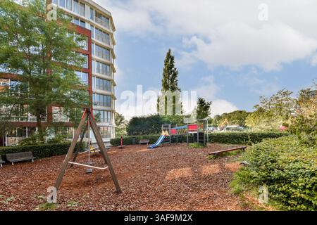 Ein lebendiger Spielplatz mit Schaukeln und Rutschen inmitten einer üppigen grünen Umgebung, die von modernen Gebäuden unter hellem Himmel umgeben ist. Stockfoto