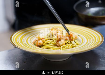 Ein Küchenchef legt gegrillte Garnelen vorsichtig mit einer Metallzange auf einen Teller Spaghetti aglio e olio. Die Pasta, die mit Olivenöl glänzt, wird garniert Stockfoto