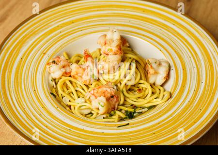Ein wunderschön überzogener Garnelen-Spaghetti-aglio e olio wird auf einer gelb gestreiften Keramikschale auf einem Holztisch präsentiert. Die Teigwaren sind mit Olivenöl überzogen Stockfoto