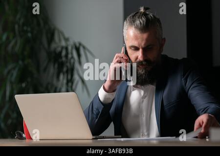 Bärtiger Geschäftsmann oder CFO in einem Anzug mit einem Smartphone am Schreibtisch mit Laptop und roter Kaffeetasse. Konzept moderner Unternehmergeist, Fernarbeit Stockfoto