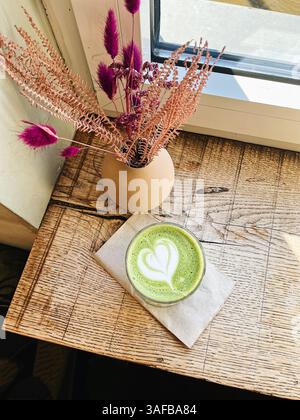 Flacher Schuss eines Matcha-flachen weiß mit Herz-Latte-Kunst auf einem rustikalen Holztisch und braune Serviette, rosa getrocknete Blumen auf einer Vase Stockfoto