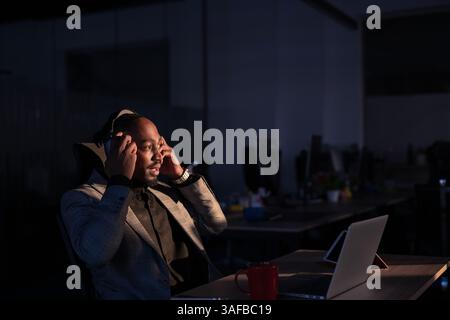Afrikanischer Geschäftsmann, der Kopfhörer trägt, Musik oder Audioinhalte hört, während er bis spät in einem modernen Büro arbeitet. Konzept von Fokus, Entspannung Stockfoto