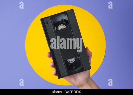 Weibliche Hand, die Videokassette auf violettem Hintergrund mit gelbem Kreis hält Stockfoto