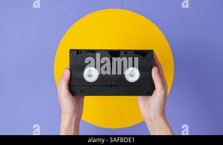 Weibliche Hand, die Videokassette auf violettem Hintergrund mit gelbem Kreis hält Stockfoto