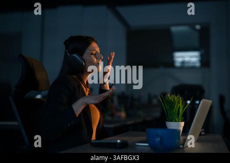 Glückliche junge Frau in einem modernen Büro, die Musik über Kopfhörer genießt, während sie lange an einem Laptop arbeitet. Konzept der Work-Life-Balance, Entspannung im The Stockfoto