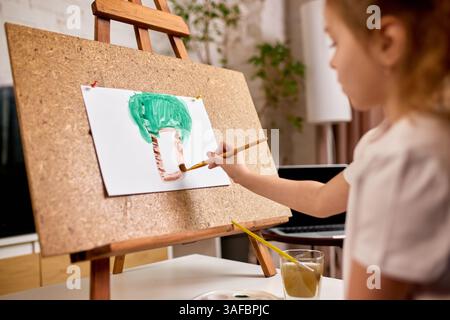 Mädchen, das zu Hause im Wohnzimmer sitzt, mit natürlichem Blitz, malte Palmen, beschäftigte sich mit Kunst in ruhiger Umgebung. Stockfoto